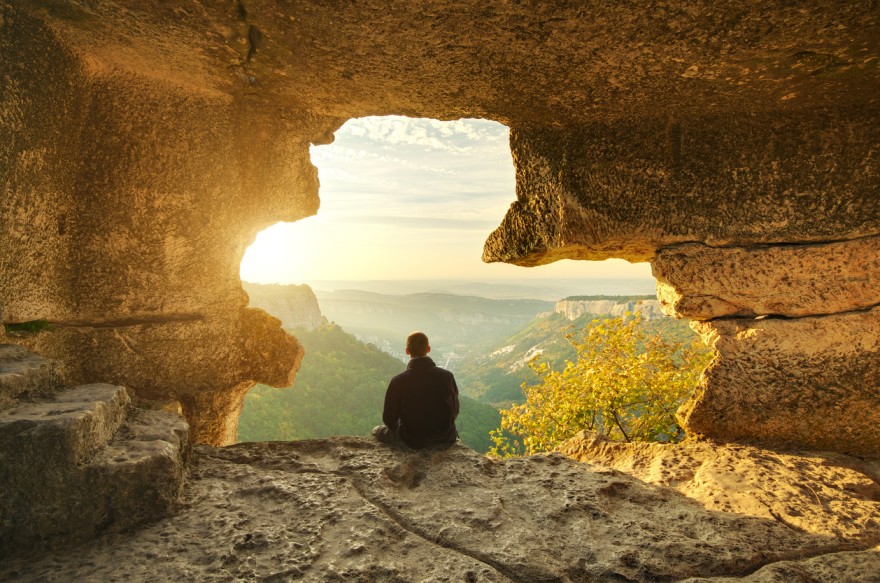 A man sitting at the edge of a cave during sunset, embodying the essence of being more and doing less 