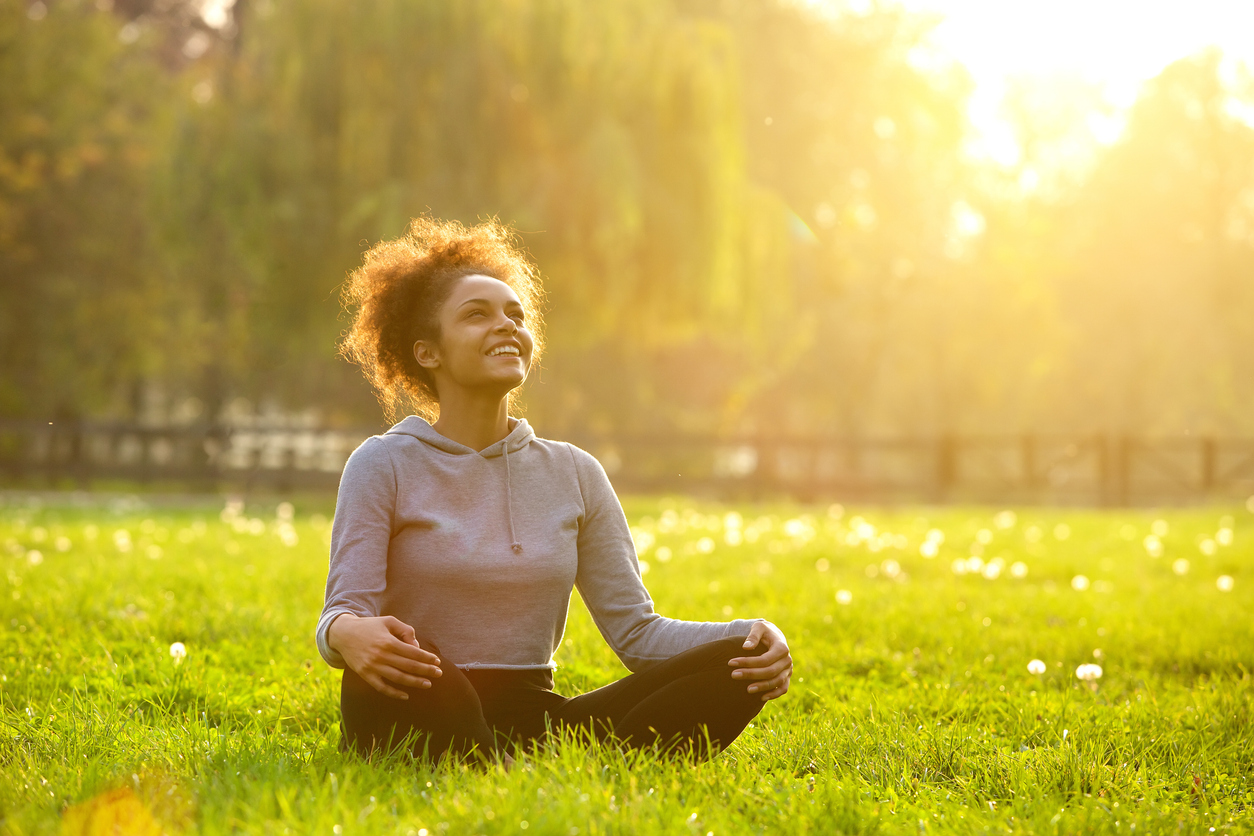 A young woman smiling contentedly with her eyes closed and face turned towards the sunlight, embodying the balance and harmony of holistic coaching in nature