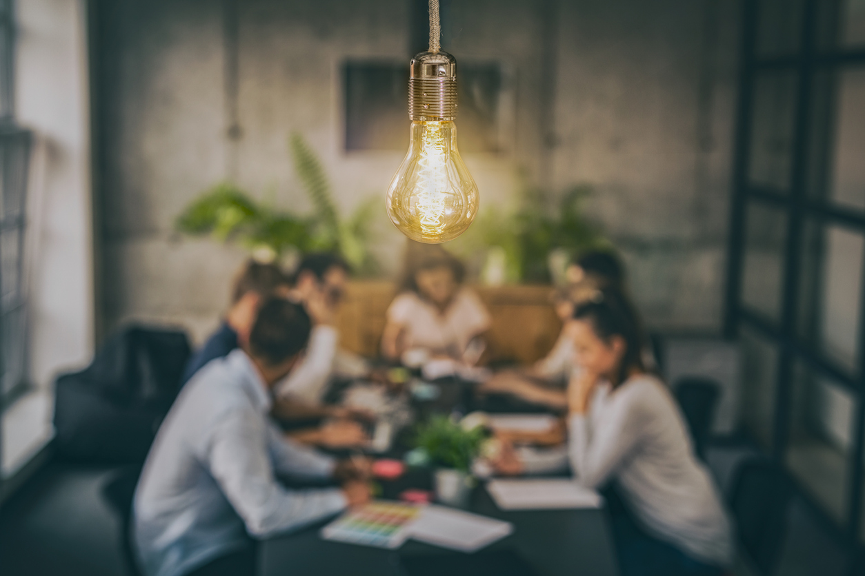 A brightly lit lightbulb hangs over a group of focused professionals, symbolizing relational leadership in the workplace.