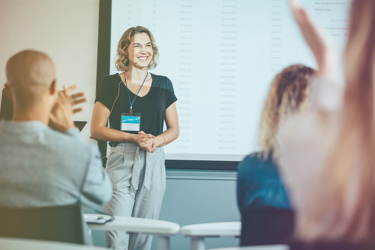 A confident woman stands at the front of a classroom as she receives applause from the audience.