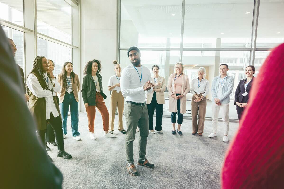 A group of business professionals are standing in a circle around a man who is confidently leading the group through positive and productive coaching.