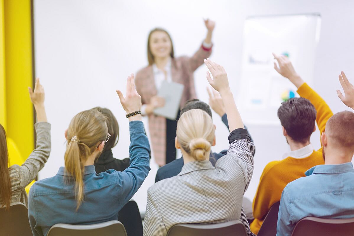 A confident female leader raises her hand and smiles as the audience raises their hands back in a sign of affirmation.