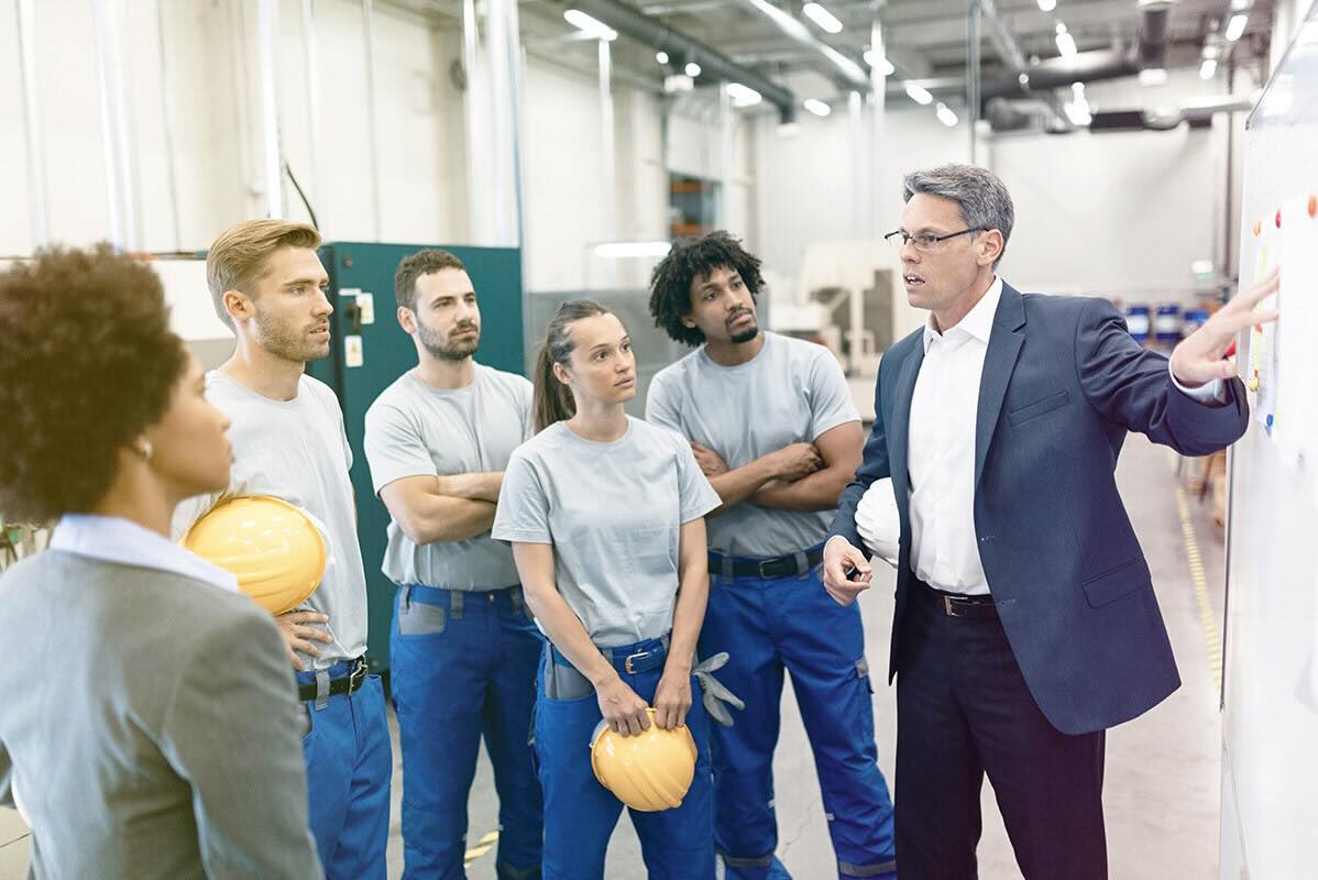 A group of employees in a factory are meeting around a large white board and are being led by a confident man in a suit holding a white hard hat.