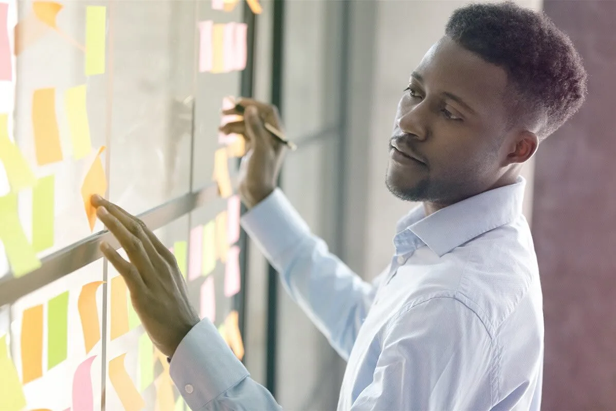 A young business professional standing in front of windows in a large office, uses sticky notes to organize his thoughts and prioritize ideas.