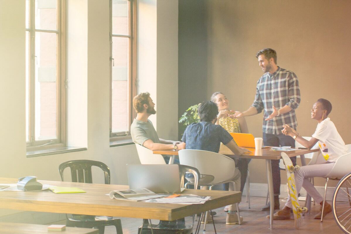 A group of five adults gather around a table in a community space, happily interacting as one individual stands and leads the conversation.