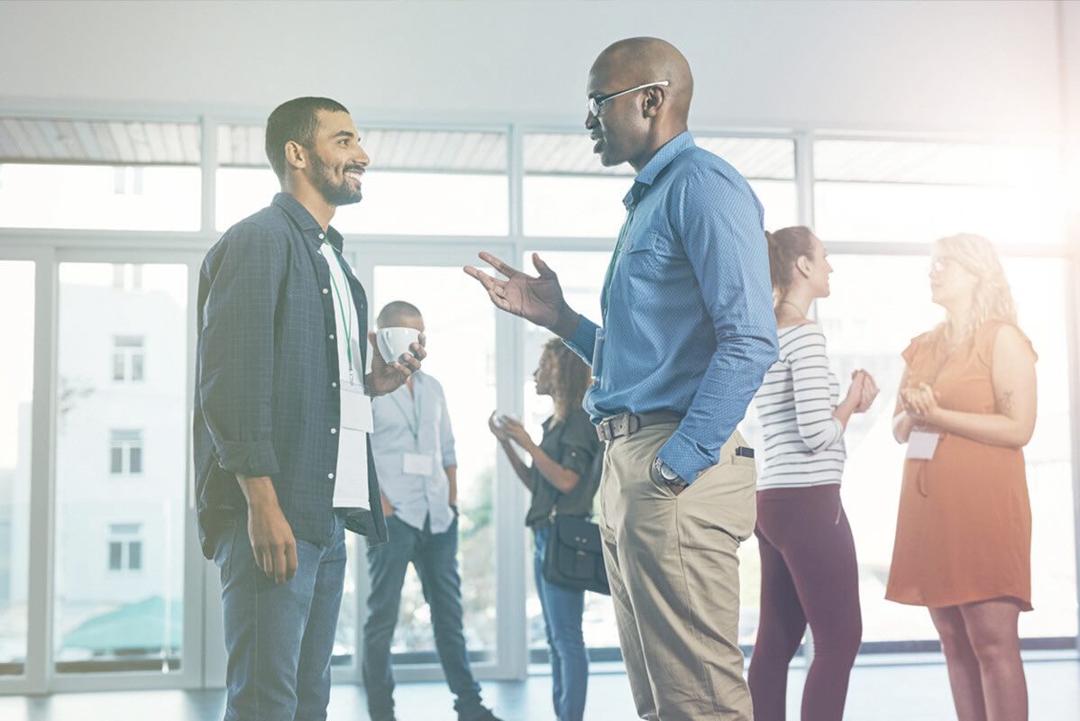 Two men engaged in professional conversation at a business-casual event with other business people mingling in the background.