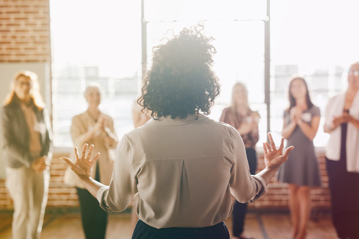 A professional woman confidently speaking to a group of women in a community meeting space.