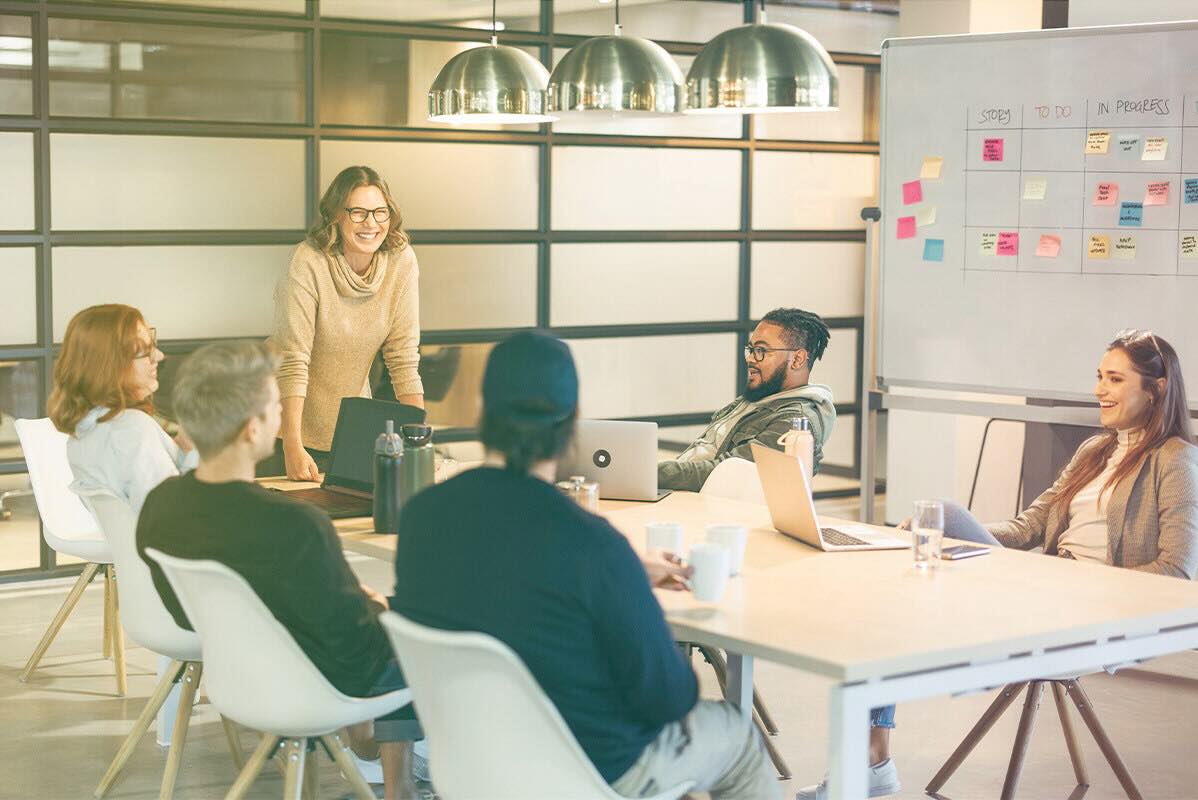 A group of six business professionals are meeting around a conference room table in an open office with laptops, coffee, and water bottles on the table.