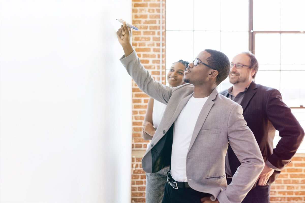 A man writes on a whiteboard while his peers watch and smile.