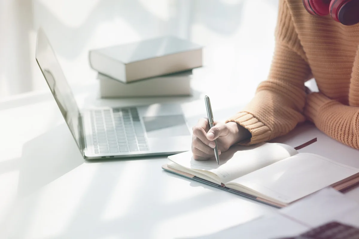 Person writing in notebook beside laptop and stacked books Person writing in notebook beside laptop and stacked books