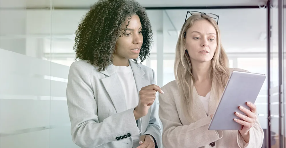 two female office workers discuss while looking at tablet