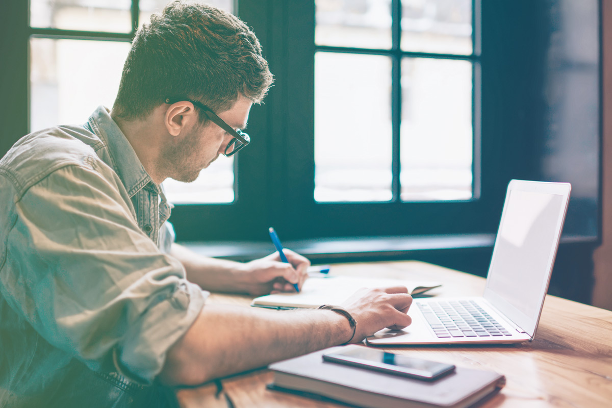 Gentleman in grey shirt writing notes and has laptop on a table