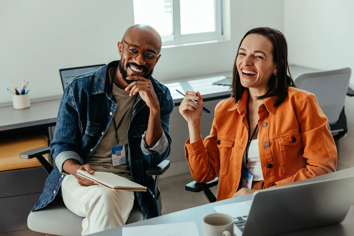 Two colleagues smiling during a workplace coaching session with laptops and notebooks.