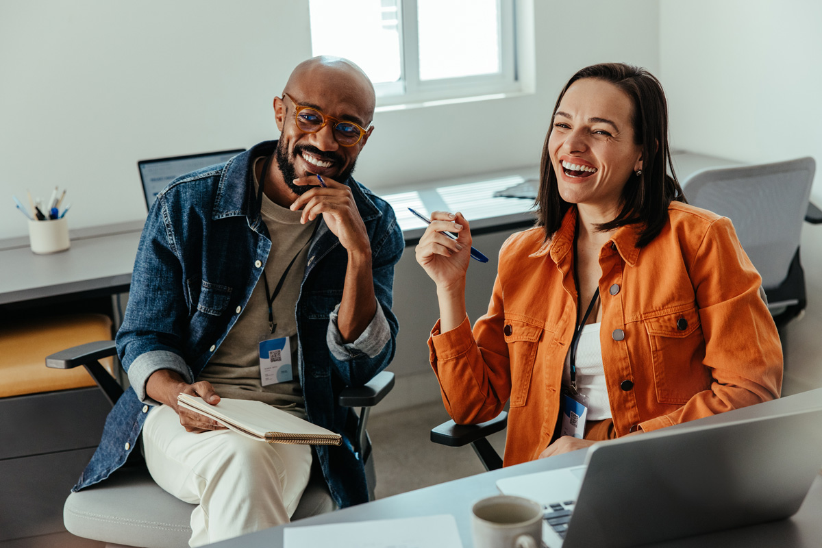 Two colleagues smiling during a workplace coaching session with laptops and notebooks.