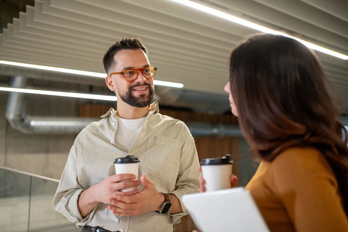 Two coworkers talking while holding coffee cups in an office.