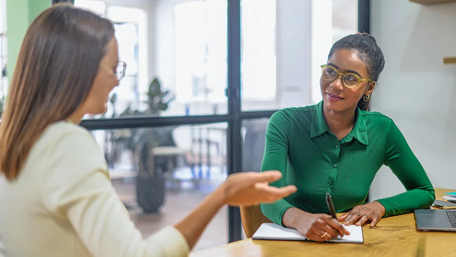 Two women talking at desk and taking notes.