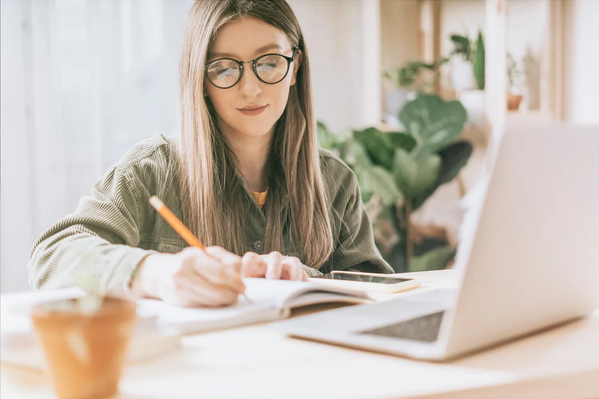 Woman writing in a notebook beside a laptop.