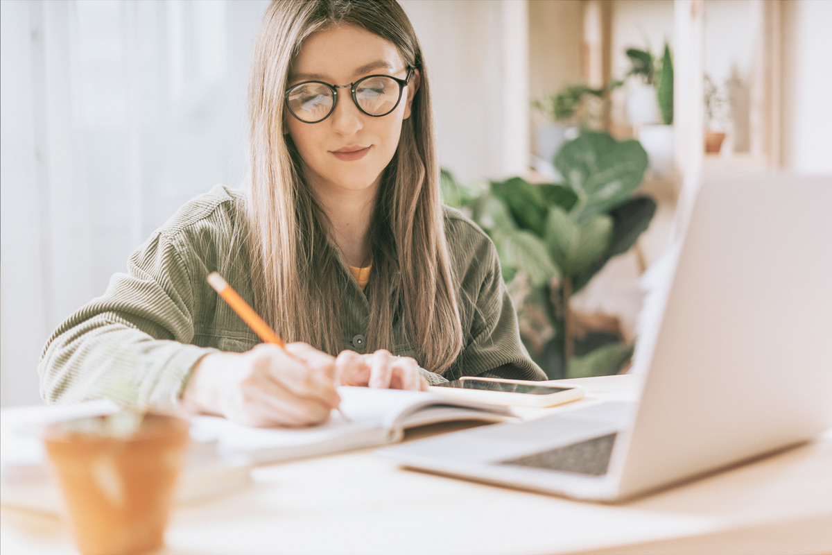 Woman writing in a notebook beside a laptop.